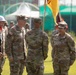 A military ceremony is taking place outdoors on a grass field, likely a change of command or unit activation ceremony. Several U.S. Army soldiers in OCP (Operational Camouflage Pattern) uniforms are gathered, with one soldier holding a yellow guidon flag. A ceremonial guard unit wearing white helmets and carrying rifles is visible in the background.