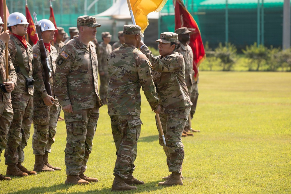 A military ceremony is taking place on a grass field, appearing to be a change of command or unit activation ceremony. A female soldier is handing or receiving a yellow guidon (unit flag) to/from another soldier, while a senior officer stands witness. Troops in OCP (Operational Camouflage Pattern) uniforms are formed in ranks in the background, with color guard members holding red and yellow flags.