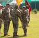 A military ceremony is taking place on a grass field, appearing to be a change of command or unit activation ceremony. A female soldier is handing or receiving a yellow guidon (unit flag) to/from another soldier, while a senior officer stands witness. Troops in OCP (Operational Camouflage Pattern) uniforms are formed in ranks in the background, with color guard members holding red and yellow flags.