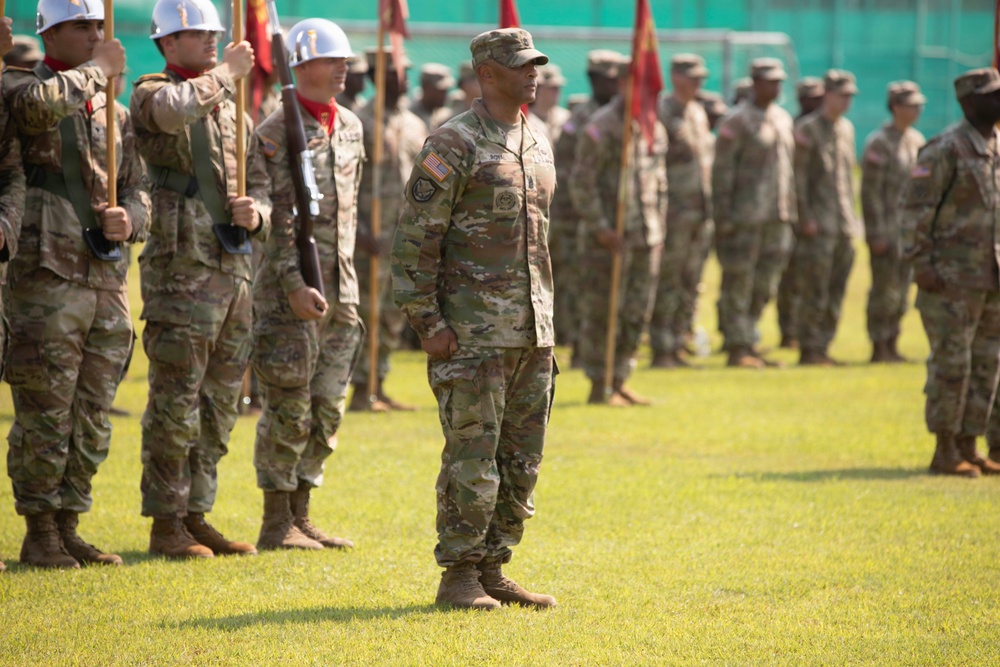 A military formation ceremony is taking place on a grass field, with soldiers in US Army OCP (Operational Camouflage Pattern) uniforms standing at attention in organized ranks. A senior soldier, likely an officer or senior NCO, stands prominently in the foreground facing the formation. Color guard members with guidons and rifles wearing white helmets are visible on the left side.