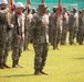 A military formation ceremony is taking place on a grass field, with soldiers in US Army OCP (Operational Camouflage Pattern) uniforms standing at attention in organized ranks. A senior soldier, likely an officer or senior NCO, stands prominently in the foreground facing the formation. Color guard members with guidons and rifles wearing white helmets are visible on the left side.
