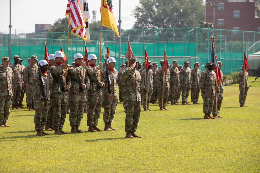 A formal military ceremony is taking place on a grass parade field, featuring soldiers in camouflage uniforms standing in formation. A color guard unit is prominently displayed in the foreground, carrying multiple flags including the United States flag, the South Korean flag, and what appear to be unit guidons. The setting appears to be a military installation, likely in South Korea based on the visible flags.