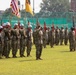 A formal military ceremony is taking place on a grass parade field, featuring soldiers in camouflage uniforms standing in formation. A color guard unit is prominently displayed in the foreground, carrying multiple flags including the United States flag, the South Korean flag, and what appear to be unit guidons. The setting appears to be a military installation, likely in South Korea based on the visible flags.