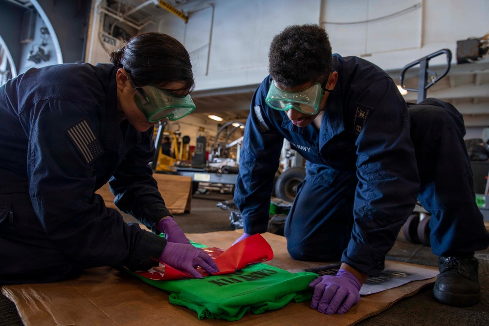 Two U.S. Navy personnel in dark blue coveralls are kneeling on the deck of what appears to be a naval vessel's hangar bay, working with green and red fabric or jersey material. Both individuals are wearing protective goggles and purple nitrile gloves, suggesting they are handling materials that require safety precautions. The background shows a large industrial hangar space with aircraft maintenance equipment and machinery visible.