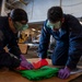 Two U.S. Navy personnel in dark blue coveralls are kneeling on the deck of what appears to be a naval vessel's hangar bay, working with green and red fabric or jersey material. Both individuals are wearing protective goggles and purple nitrile gloves, suggesting they are handling materials that require safety precautions. The background shows a large industrial hangar space with aircraft maintenance equipment and machinery visible.