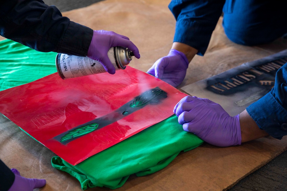 Two individuals wearing purple latex gloves are applying spray paint through a stencil onto what appears to be a green garment or fabric. A red base layer is visible beneath a dark stencil, with black and green spray paint being actively applied. A second stencil with text is visible on the brown paper surface to the right.