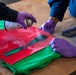 Two individuals wearing purple latex gloves are applying spray paint through a stencil onto what appears to be a green garment or fabric. A red base layer is visible beneath a dark stencil, with black and green spray paint being actively applied. A second stencil with text is visible on the brown paper surface to the right.