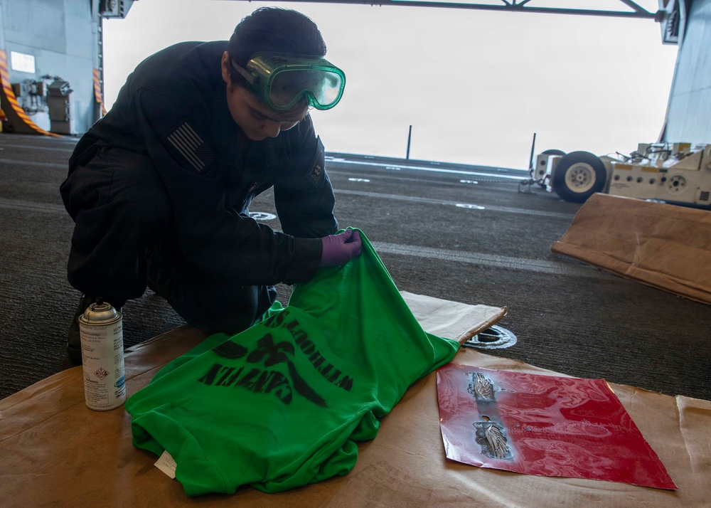 A military service member wearing green safety goggles and purple nitrile gloves is spray-painting a stencil design onto a green garment, likely a shirt or vest. The work is being done in what appears to be a hangar bay of a naval vessel, with aircraft handling equipment visible in the background. A spray paint can and a red stencil sheet with figure designs are also present on the work surface.