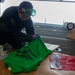 A military service member wearing green safety goggles and purple nitrile gloves is spray-painting a stencil design onto a green garment, likely a shirt or vest. The work is being done in what appears to be a hangar bay of a naval vessel, with aircraft handling equipment visible in the background. A spray paint can and a red stencil sheet with figure designs are also present on the work surface.