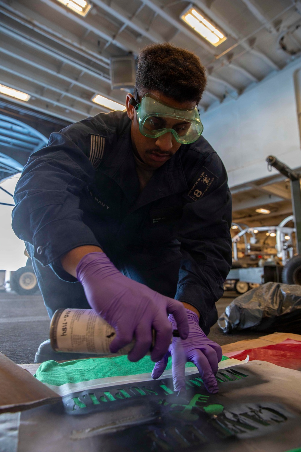 A U.S. Navy sailor wearing protective goggles and purple nitrile gloves is working on a surface, applying or cleaning something using a cloth and a canister product. The work is being performed in what appears to be a hangar bay of a naval vessel, with equipment and machinery visible in the background. The sailor is wearing a dark Navy coverall uniform with a U.S. flag patch and unit insignia.