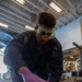 A U.S. Navy sailor wearing protective goggles and purple nitrile gloves is working on a surface, applying or cleaning something using a cloth and a canister product. The work is being performed in what appears to be a hangar bay of a naval vessel, with equipment and machinery visible in the background. The sailor is wearing a dark Navy coverall uniform with a U.S. flag patch and unit insignia.