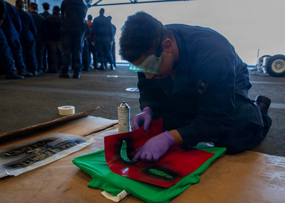 A person wearing blue coveralls, purple nitrile gloves, and green safety goggles is kneeling on the floor applying a stencil design onto a green t-shirt using spray paint. A red stencil sheet is placed over the shirt, and a spray paint can is visible nearby. In the background, a group of people in similar dark uniforms are gathered, suggesting a military or organized group setting.