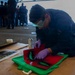 A person wearing blue coveralls, purple nitrile gloves, and green safety goggles is kneeling on the floor applying a stencil design onto a green t-shirt using spray paint. A red stencil sheet is placed over the shirt, and a spray paint can is visible nearby. In the background, a group of people in similar dark uniforms are gathered, suggesting a military or organized group setting.