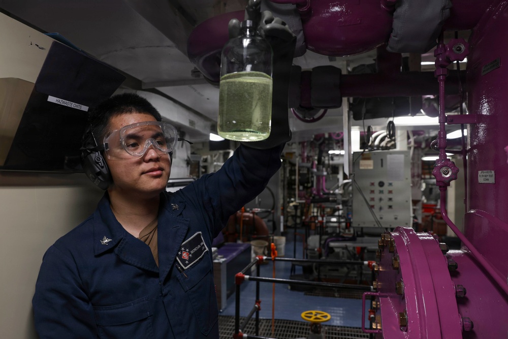 A U.S. Navy sailor in a dark blue uniform is holding up a glass jar containing a yellowish-green liquid for inspection inside what appears to be a ship's engineering or machinery space. The sailor is wearing safety goggles and hearing protection, suggesting a hazardous or loud work environment. The background reveals complex industrial piping, valves, and control panels painted in purple/magenta, characteristic of a naval vessel's interior.