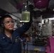 A U.S. Navy sailor in a dark blue uniform is holding up a glass jar containing a yellowish-green liquid for inspection inside what appears to be a ship's engineering or machinery space. The sailor is wearing safety goggles and hearing protection, suggesting a hazardous or loud work environment. The background reveals complex industrial piping, valves, and control panels painted in purple/magenta, characteristic of a naval vessel's interior.