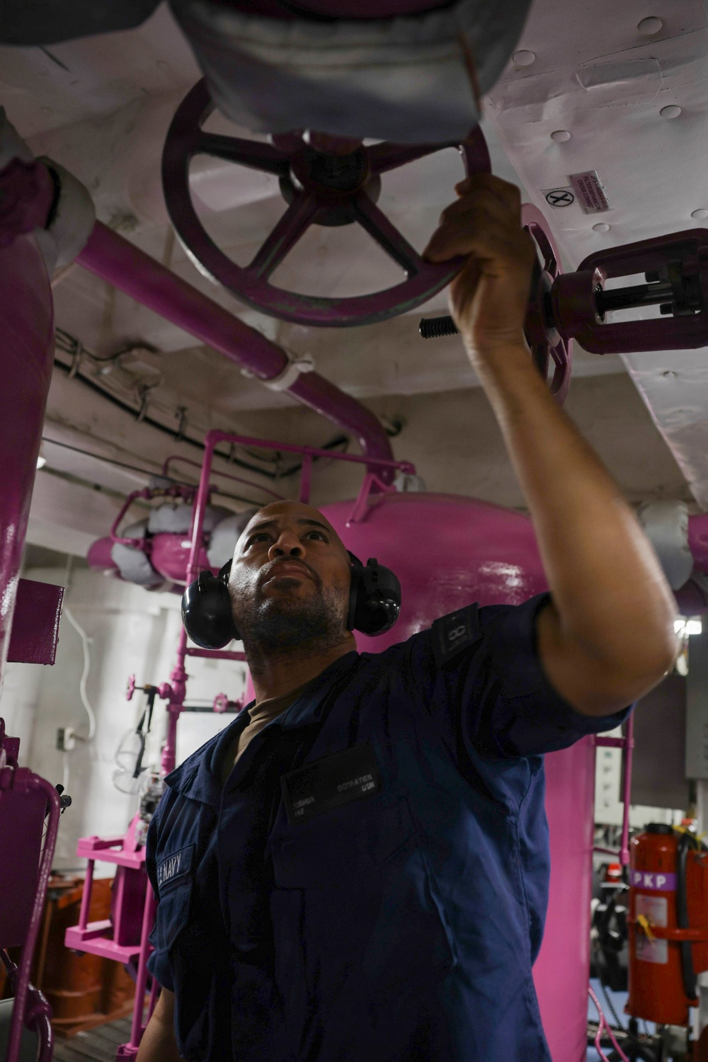 A U.S. Navy sailor wearing hearing protection is operating or inspecting an overhead valve wheel mechanism in what appears to be a ship's engineering or machinery space. The environment is characterized by bright pink/magenta painted pipes and equipment, which is consistent with aviation fuel (JP-5) piping color coding used on U.S. Navy vessels. The sailor is dressed in Navy blue coveralls and is reaching upward to manipulate the valve.
