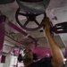 A U.S. Navy sailor wearing hearing protection is operating or inspecting an overhead valve wheel mechanism in what appears to be a ship's engineering or machinery space. The environment is characterized by bright pink/magenta painted pipes and equipment, which is consistent with aviation fuel (JP-5) piping color coding used on U.S. Navy vessels. The sailor is dressed in Navy blue coveralls and is reaching upward to manipulate the valve.