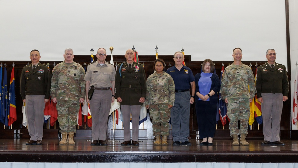 A formal group photograph of eight individuals standing on a stage in front of a display of military unit flags and guidons. The group consists of six individuals in U.S. Army military uniforms (OCP/ACU pattern and dress greens) and two civilians in business casual attire. This appears to be an official military ceremony or recognition event.
