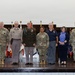 A formal group photograph of eight individuals standing on a stage in front of a display of military unit flags and guidons. The group consists of six individuals in U.S. Army military uniforms (OCP/ACU pattern and dress greens) and two civilians in business casual attire. This appears to be an official military ceremony or recognition event.