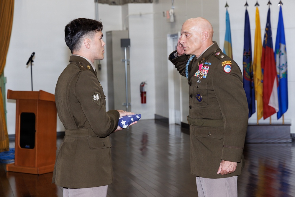 Two U.S. Army soldiers are participating in a formal military ceremony indoors. A junior enlisted soldier is presenting a folded American flag to a senior officer who is saluting. The senior officer is wearing numerous medals and decorations, including what appears to be a blue medal around his neck, suggesting a retirement or awards ceremony.