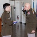 Two U.S. Army soldiers are participating in a formal military ceremony indoors. A junior enlisted soldier is presenting a folded American flag to a senior officer who is saluting. The senior officer is wearing numerous medals and decorations, including what appears to be a blue medal around his neck, suggesting a retirement or awards ceremony.