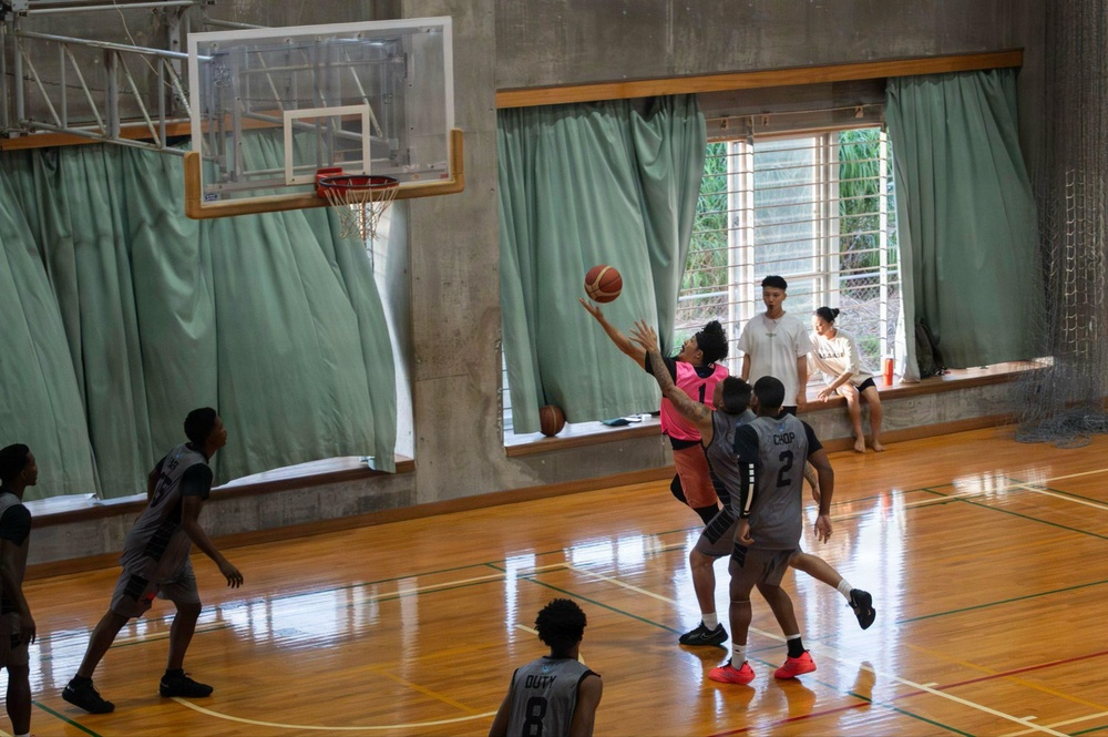 An indoor basketball game is being played in a gymnasium with a wooden court floor. A player in a pink jersey (number 1) is attempting a layup near the basket while being defended by players in gray uniforms. Two spectators are seated on a windowsill in the background watching the game.