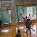 An indoor basketball game is being played in a gymnasium with a wooden court floor. A player in a pink jersey (number 1) is attempting a layup near the basket while being defended by players in gray uniforms. Two spectators are seated on a windowsill in the background watching the game.