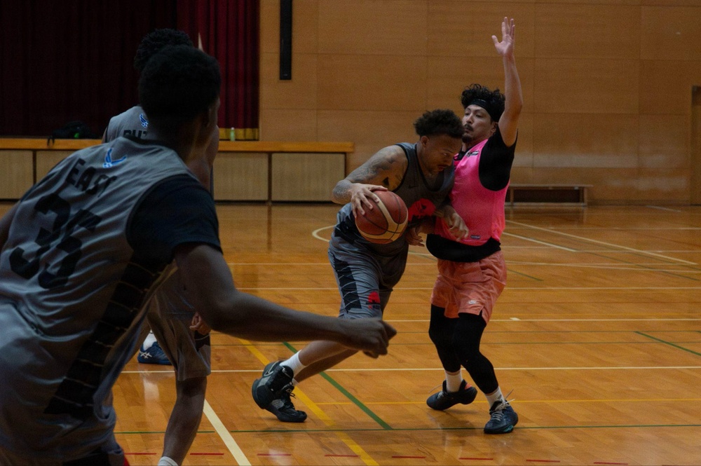 A basketball game or practice session is taking place on an indoor hardwood court. A player in a gray uniform is driving with the ball while being defended by a player wearing a pink vest/pinnie over a black outfit. Additional players in gray uniforms are visible in the foreground and background.