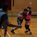 A basketball game or practice session is taking place on an indoor hardwood court. A player in a gray uniform is driving with the ball while being defended by a player wearing a pink vest/pinnie over a black outfit. Additional players in gray uniforms are visible in the foreground and background.