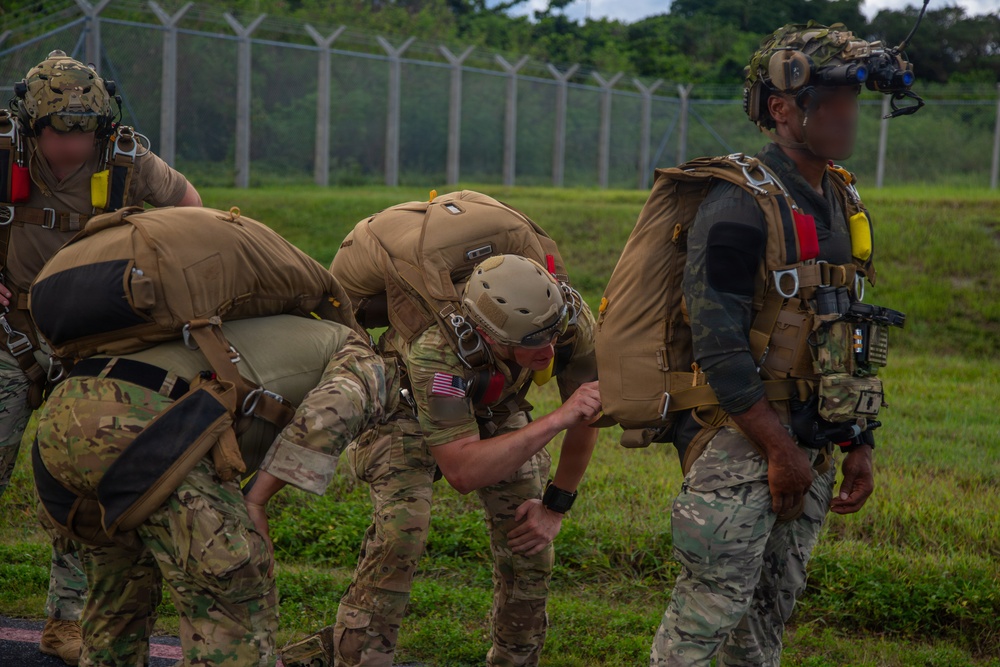 A group of military personnel in full tactical gear, including parachute rigs and helmets with night vision mounts, are preparing or conducting post-jump activities on a grassy area near a perimeter fence. At least one soldier displays a US flag patch on their sleeve, indicating US military affiliation. The personnel appear to be adjusting or checking their equipment in what looks like a pre- or post-parachute jump scenario.