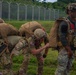 A group of military personnel in full tactical gear, including parachute rigs and helmets with night vision mounts, are preparing or conducting post-jump activities on a grassy area near a perimeter fence. At least one soldier displays a US flag patch on their sleeve, indicating US military affiliation. The personnel appear to be adjusting or checking their equipment in what looks like a pre- or post-parachute jump scenario.