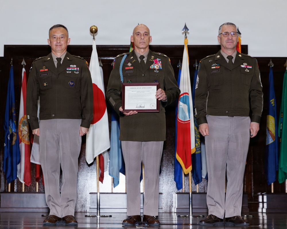 Three U.S. Army soldiers in olive green service uniforms are posed for a formal military ceremony photograph. The center figure is holding a framed 'Certificate of Appreciation' and displays numerous medals and a distinctive blue aiguillette. They are standing in front of a display of multiple flags including the Japanese national flag and various U.S. state flags.