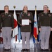 Three U.S. Army soldiers in olive green service uniforms are posed for a formal military ceremony photograph. The center figure is holding a framed 'Certificate of Appreciation' and displays numerous medals and a distinctive blue aiguillette. They are standing in front of a display of multiple flags including the Japanese national flag and various U.S. state flags.
