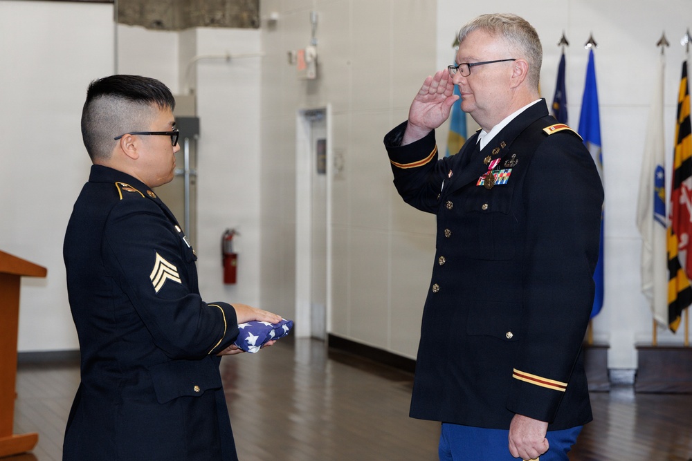Two U.S. Army soldiers in dress uniforms are participating in a formal military ceremony indoors. The senior officer on the right is rendering a salute while the soldier on the left holds a folded American flag. State and military flags are visible in the background, suggesting an official ceremony such as a retirement, promotion, or memorial event.