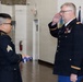 Two U.S. Army soldiers in dress uniforms are participating in a formal military ceremony indoors. The senior officer on the right is rendering a salute while the soldier on the left holds a folded American flag. State and military flags are visible in the background, suggesting an official ceremony such as a retirement, promotion, or memorial event.