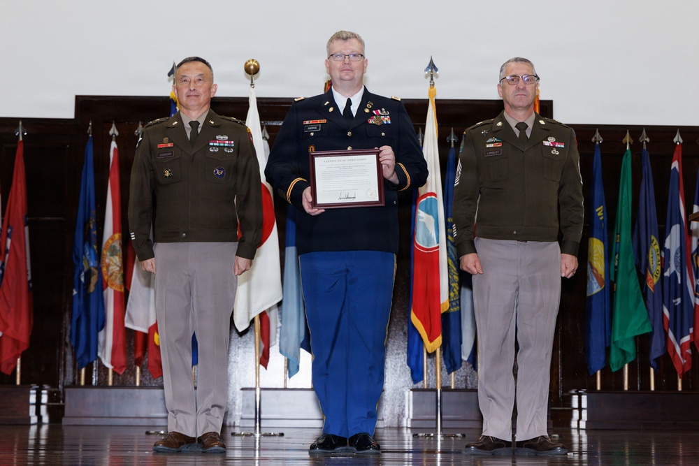 Three U.S. Army personnel are posed in a formal military ceremony, with the center individual holding a framed certificate or award. The two flanking officers wear the Army Green Service Uniform (OCP-based greens), while the center individual wears the Army Service Uniform (ASU) in blue. Multiple national and state flags are displayed in the background, indicating a formal headquarters or ceremonial hall setting.