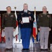 Three U.S. Army personnel are posed in a formal military ceremony, with the center individual holding a framed certificate or award. The two flanking officers wear the Army Green Service Uniform (OCP-based greens), while the center individual wears the Army Service Uniform (ASU) in blue. Multiple national and state flags are displayed in the background, indicating a formal headquarters or ceremonial hall setting.