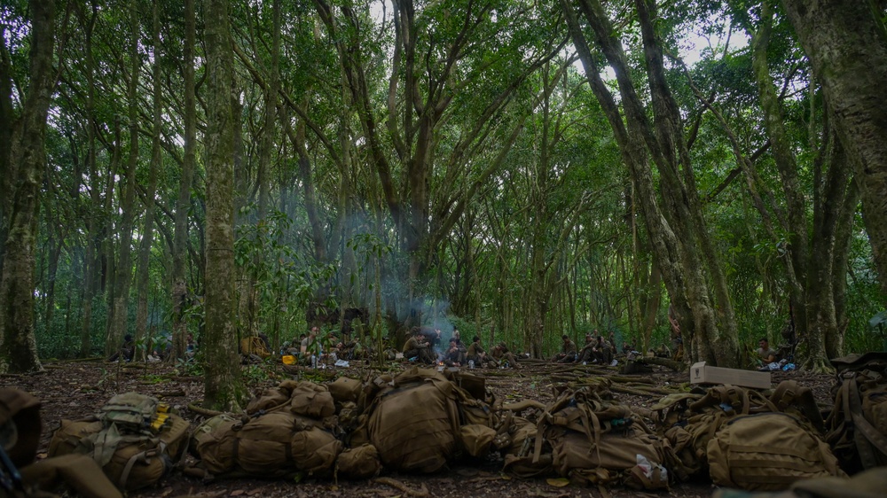 A group of military personnel are resting in a dense tropical forest clearing, with their olive-drab/tan tactical backpacks and gear piled on the ground in the foreground. A small fire or cooking source is visible in the middle distance, producing a thin column of smoke rising through the forest canopy. The scene appears to be a field halt or bivouac during a jungle training exercise or operation.