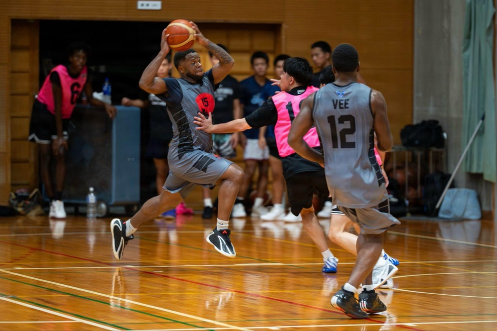 An indoor basketball game is in progress on a hardwood court, likely in an Asian gymnasium based on the architectural style. A player in a gray uniform wearing number 15 is airborne, holding the ball in a passing or shooting position while being defended by players in pink pinnies. Teammates and spectators are visible in the background.