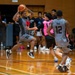 An indoor basketball game is in progress on a hardwood court, likely in an Asian gymnasium based on the architectural style. A player in a gray uniform wearing number 15 is airborne, holding the ball in a passing or shooting position while being defended by players in pink pinnies. Teammates and spectators are visible in the background.