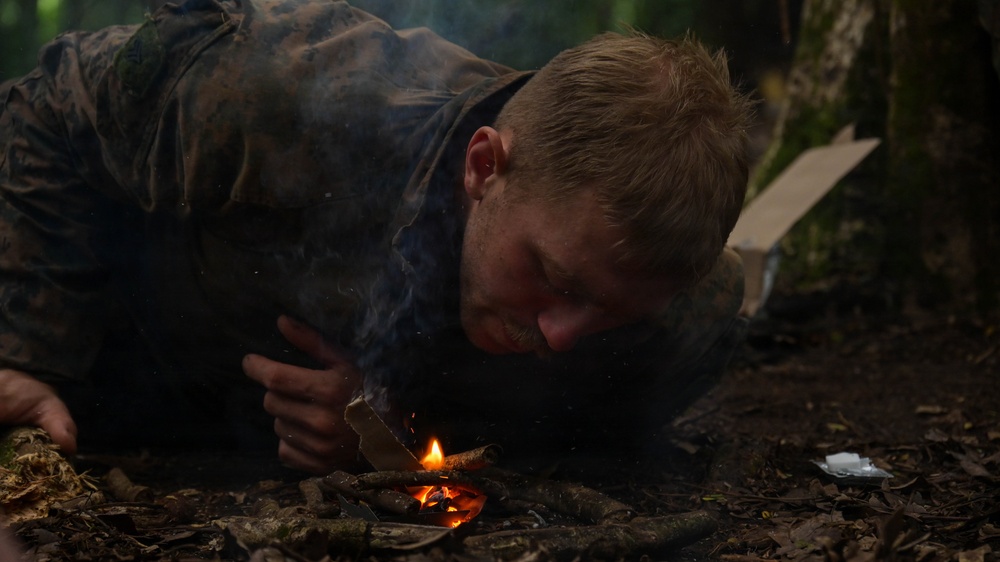 A young man wearing camouflage clothing is lying prone on a forest floor, blowing on a small fire to coax it into flame. The scene appears to depict a wilderness survival or bushcraft fire-starting exercise. Smoke rises from the small flame, and the surrounding environment is a dense, dark woodland.