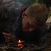 A young man wearing camouflage clothing is lying prone on a forest floor, blowing on a small fire to coax it into flame. The scene appears to depict a wilderness survival or bushcraft fire-starting exercise. Smoke rises from the small flame, and the surrounding environment is a dense, dark woodland.