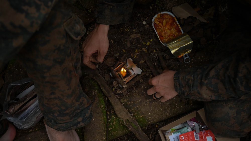 Two individuals wearing military camouflage uniforms are preparing a field meal in a dark, wooded outdoor environment. One person is lighting or tending to a small portable solid-fuel stove while the other holds an open tin can containing what appears to be a food ration. Various field ration supplies and packaging are visible nearby.