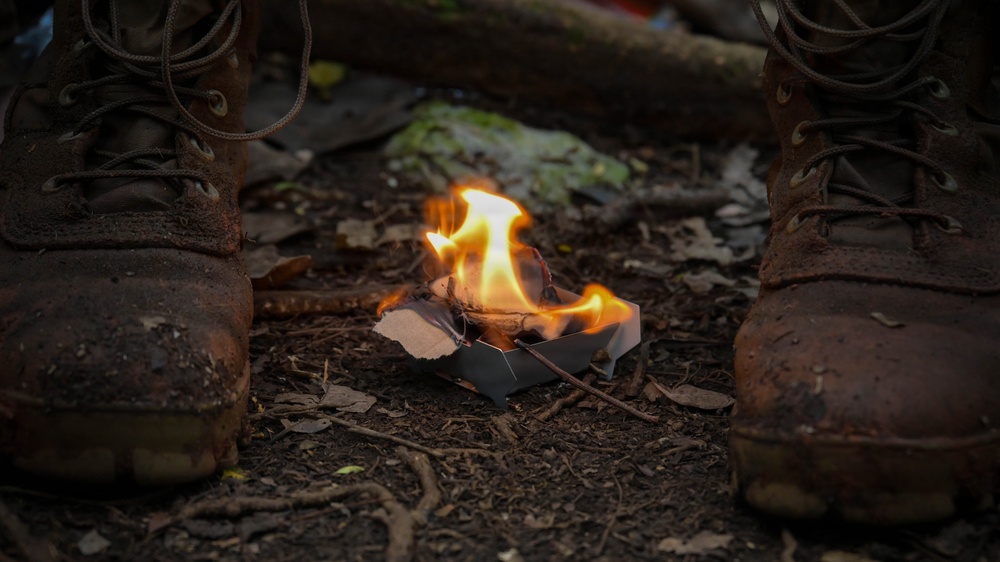 A small fire burns in a compact metal camp stove placed on forest ground between two heavily worn, mud-covered boots. The flames are fed by what appears to be cardboard and small twigs. The setting suggests an outdoor survival or bushcraft scenario.