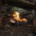 A small fire burns in a compact metal camp stove placed on forest ground between two heavily worn, mud-covered boots. The flames are fed by what appears to be cardboard and small twigs. The setting suggests an outdoor survival or bushcraft scenario.