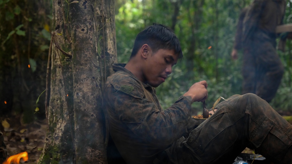 A young man in camouflage military-style clothing is seated against a large tree trunk in a dense jungle environment, eating from a small container or mess kit. There is an open fire visible in the lower left corner and smoke drifting through the scene. A second partially visible figure can be seen in the blurred background to the right.