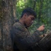 A young man in camouflage military-style clothing is seated against a large tree trunk in a dense jungle environment, eating from a small container or mess kit. There is an open fire visible in the lower left corner and smoke drifting through the scene. A second partially visible figure can be seen in the blurred background to the right.