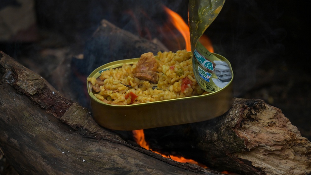 A gold/tin can containing a rice-based dish with meat and vegetables is being heated directly over an open campfire, resting between two logs. The lid of the can is peeled back and folded upward, revealing the contents inside. This appears to be an outdoor survival or camping meal preparation scenario.
