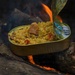 A gold/tin can containing a rice-based dish with meat and vegetables is being heated directly over an open campfire, resting between two logs. The lid of the can is peeled back and folded upward, revealing the contents inside. This appears to be an outdoor survival or camping meal preparation scenario.