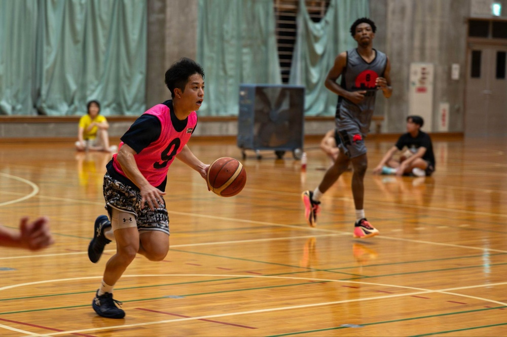 A basketball game or practice session is taking place on an indoor hardwood court. A player wearing a pink vest numbered '9' is dribbling the ball while a taller player in a gray uniform follows behind. Several spectators are seated along the sidelines in what appears to be a gymnasium.